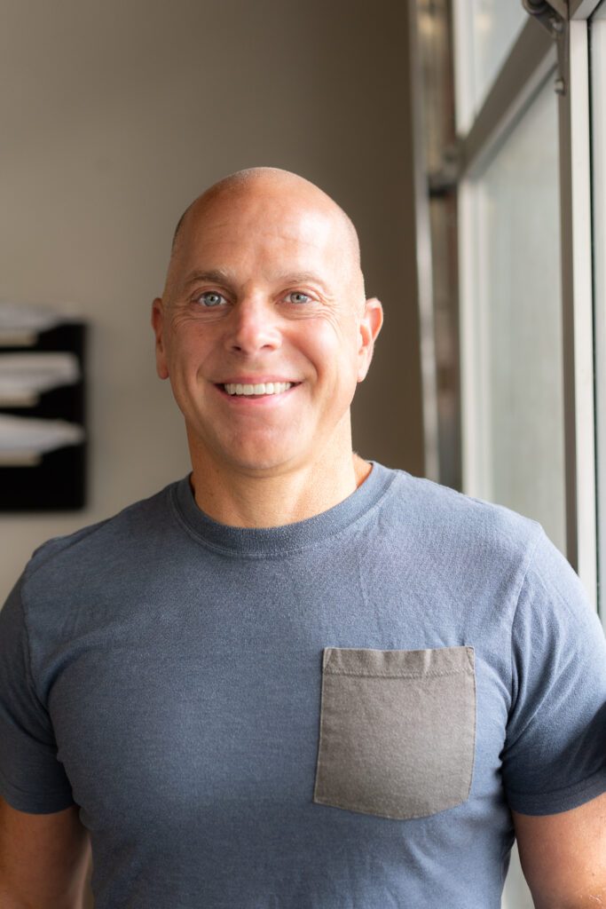 A portrait headshot of Jay Dicharry in a grey shirt smiling at the camera