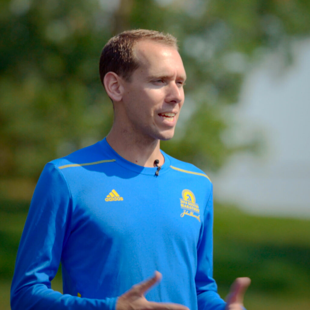 Running coach Jason Fitzgerald stands with his hands open while talking in a blue shirt with green trees in the background