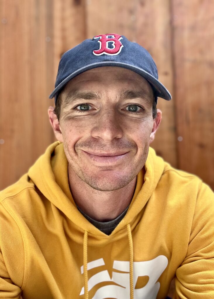 Mario Fraioli looks at the camera in a yellow hoodie and a Boston Red Sox navy cap with a closed smile