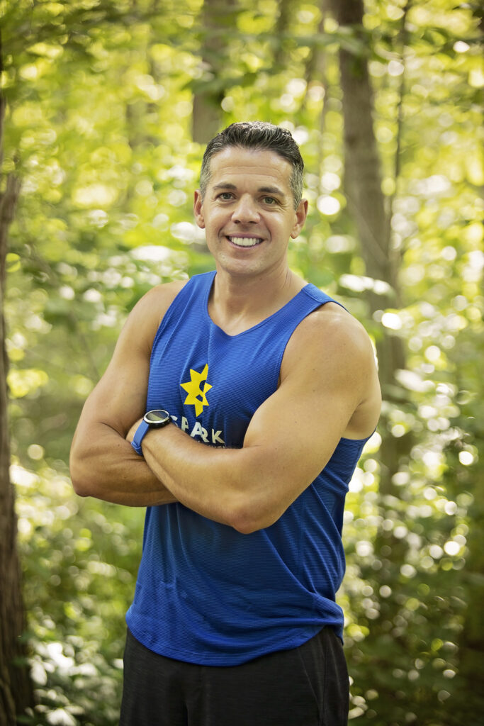 Achilles expert Duane Scotti stands with arms crossed in a blue tank top among trees